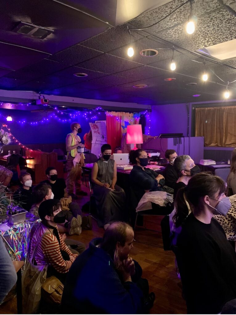 Photo of an audience. The are wearing masks with some seated in chairs while others sit on the floor. There are blue string lights on the wall in the background.
