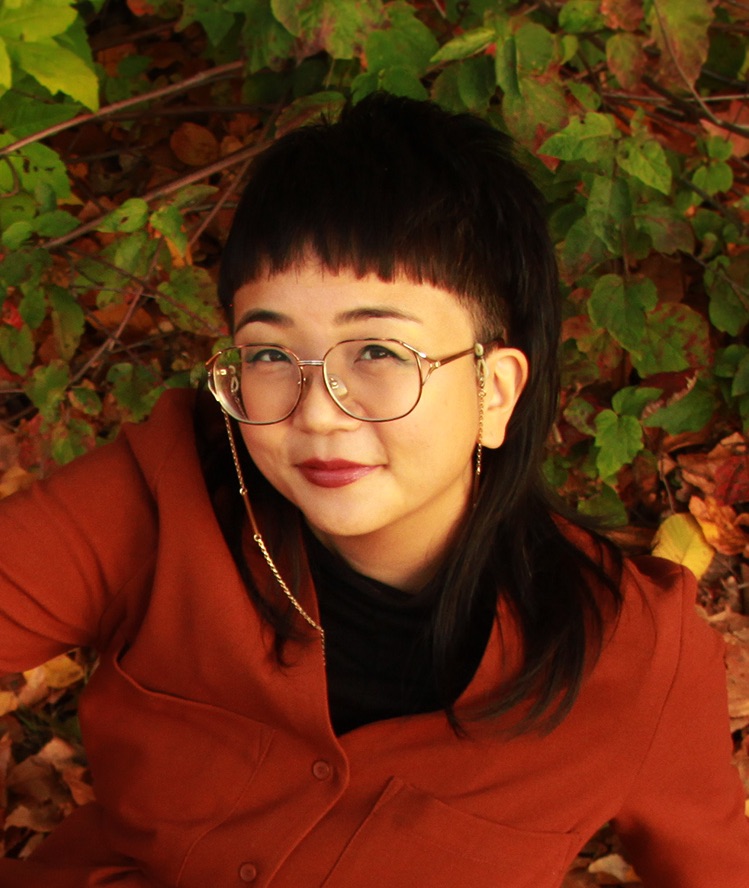Author headshot of a femme-presenting Asian person wearing a red sweater and eyeglasses with straps, sitting against a foliage background.